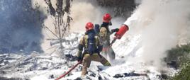Na zdjęciu widać dwóch strażaków w specjalistycznym ubraniu ochronnym, a aparatem powietrznym na plecach i maskach na twarzy, którzy gaszą pożar podając pianę gaśniczą.
