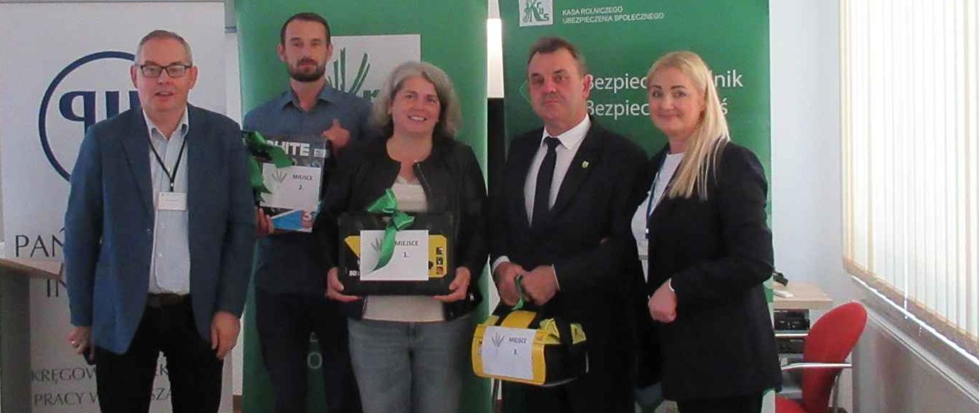 Grupa pięciu osób – dwie kobiety i trzech mężczyzn pozuje do wspólnego zdjęcia., są uśmiechnięci. Trzy osoby w środku trzymają nagrody w postaci narzędzi i elektronarzędzi, są to laureaci w konkursie testowym KRUS.