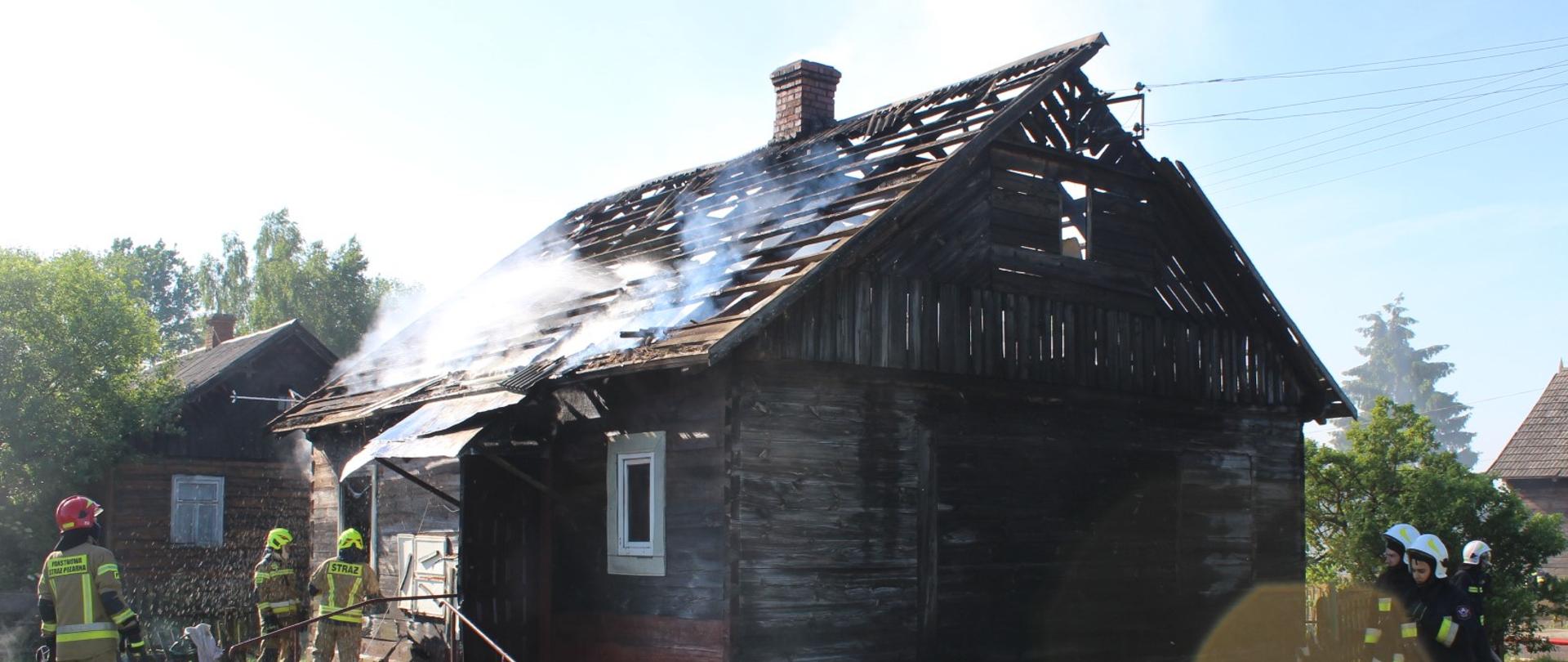 Drewniany budynek mieszkalny. Konstrukcja dachu zniszczona. Z poddasza wydobywa się dym. Przed budynkiem strażacy biorący udział w akcji gaśniczej.