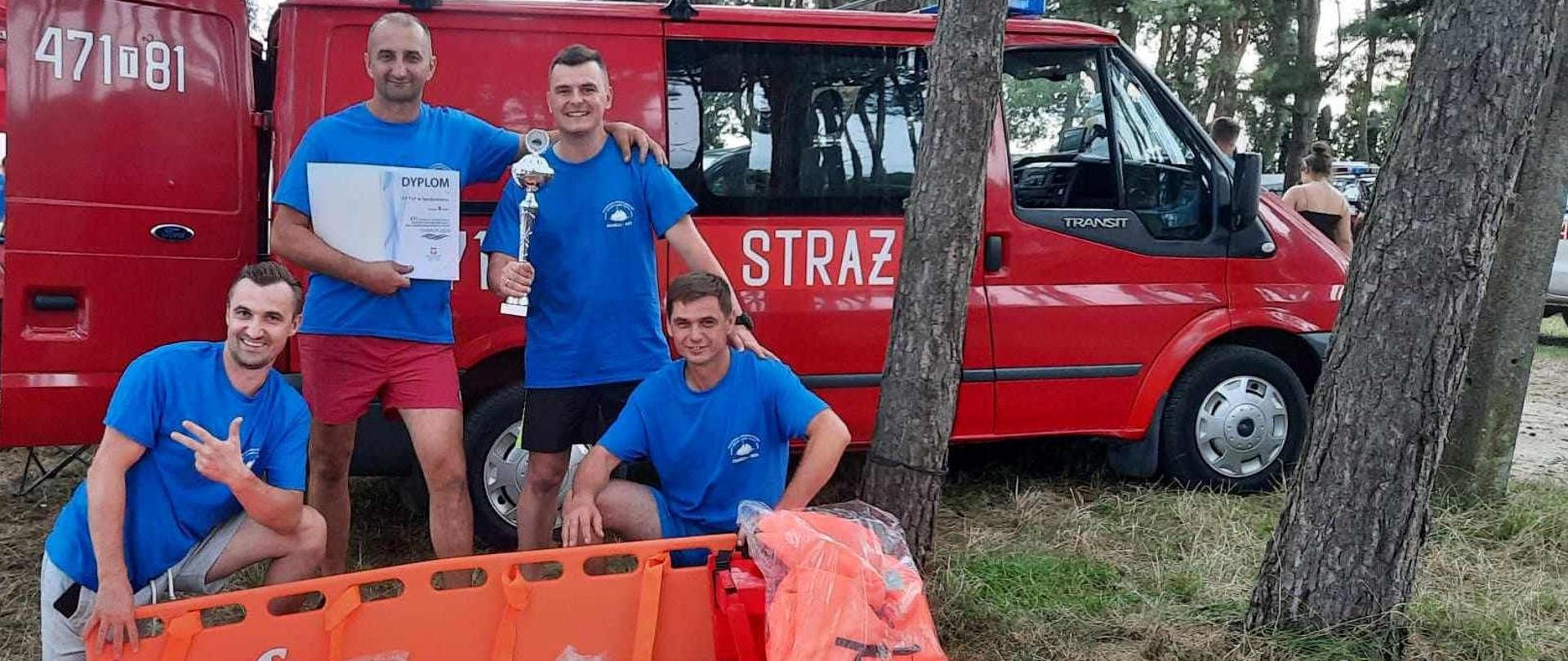 Zdjęcie zrobione w dzień nad zalewem Chańcza. Na zdjęciu widać czterech strażaków, którzy trzymają puchar, dyplom, deskę i kapoki na tle samochodu strażackiego. 