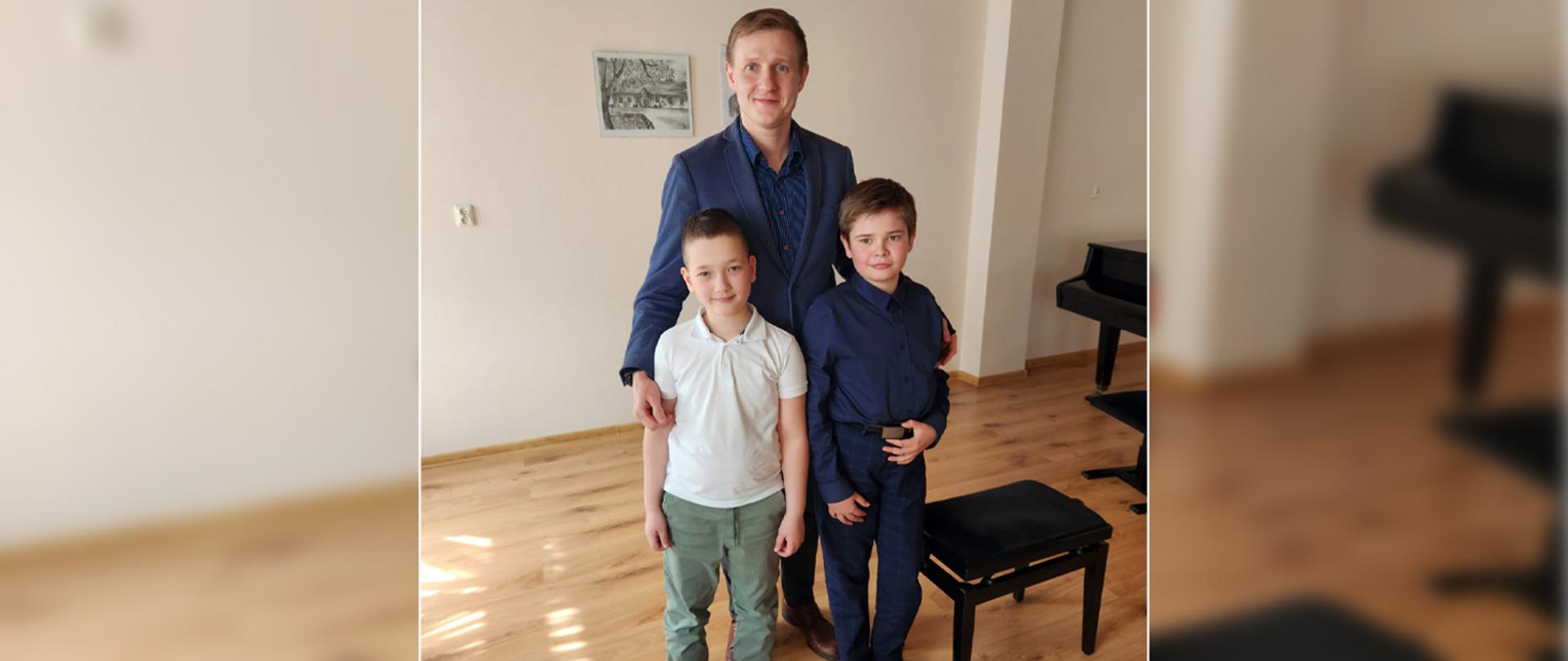 Dwoje uczniów stoi w auli wraz z nauczycielem.