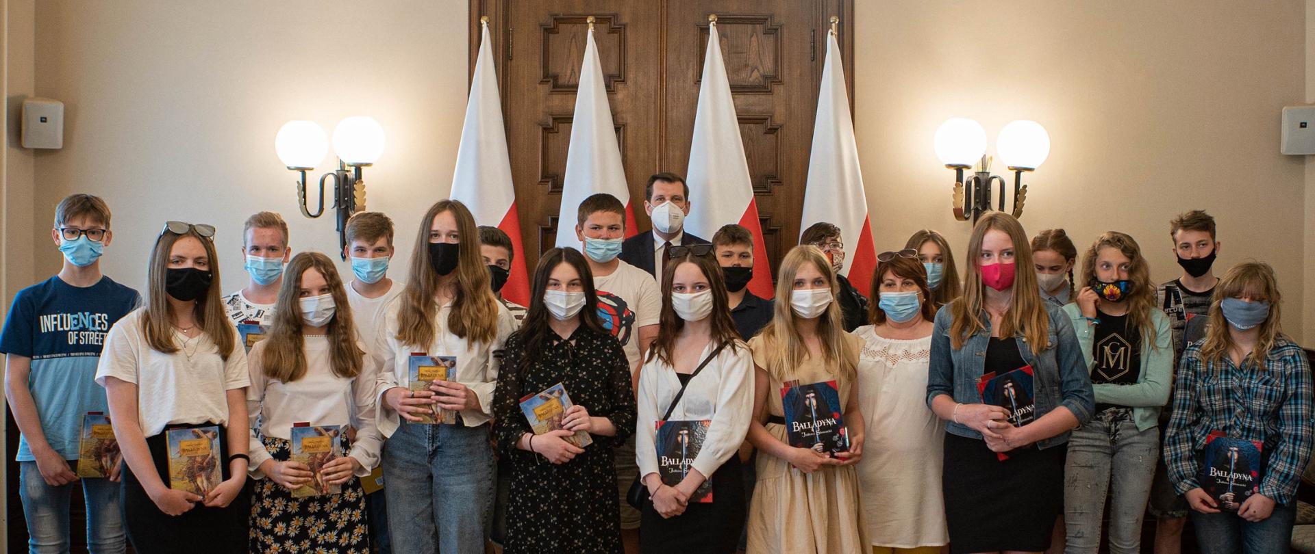 Wojewoda łódzki Tobiasz Bocheński wraz z uczniami klasy 7C Szkoły Podstawowej nr 2 w Koluszkach stoją i pozują do zdjęcia na tle flag o barwie białoczerwonej