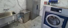 pomieszczenie gospodarcze, w którym znajdują się myjka i suszarka do masek, zlew, suszarka do ubrań, pralnica, na ścianie dozowniki płynów dezynfekujących