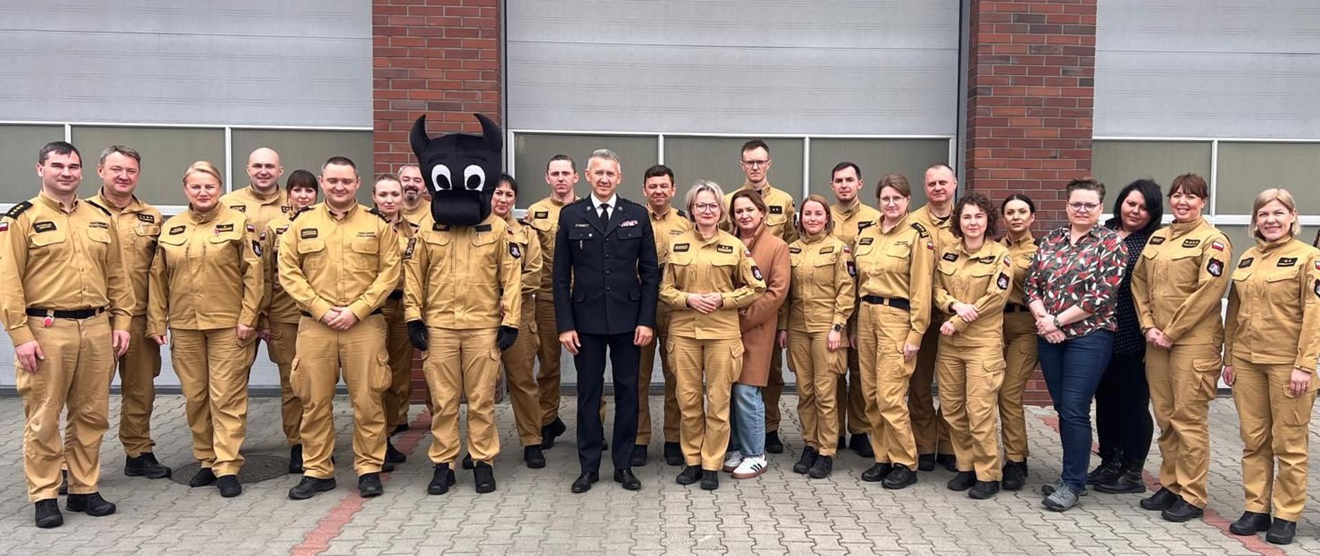 Uczestnicy spotkania PSP stoją przed garażem jednostki straży pożarnej. Wśród nich osoby w żółtych mundurach służbowych, jedna osoba w przebraniu maskotki „Bebok” oraz strażak w czarnym mundurze wyjściowym PSP, wszyscy uśmiechnięci, pozujący do zdjęcia grupowego.