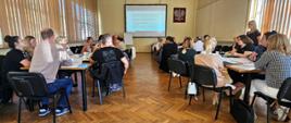 W sali konferencyjnej kilkanaście osób siedzi przy stołach, uczestnicząc w spotkaniu lub szkoleniu. Na ścianie z przodu znajduje się ekran z wyświetloną prezentacją oraz godło Polski. Uczestnicy słuchają prelegentki stojącej po prawej stronie. Pomieszczenie jest jasne, z dużymi oknami zasłoniętymi pionowymi żaluzjami.