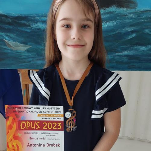 Dziewczynka stoi trzymając dyplom Brązowego Medalu w rękach, na szyi ma zawieszony brązowy medal w kształcie klucza wiolinowego.