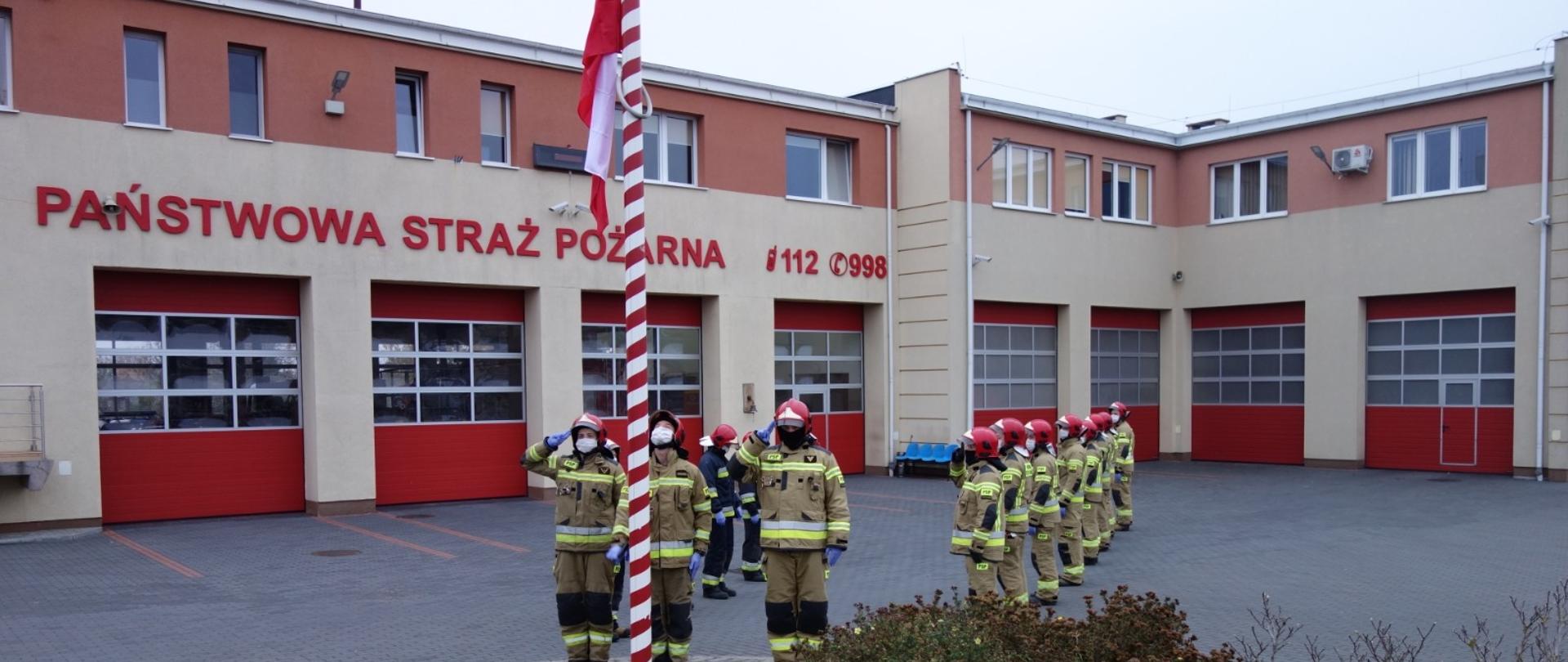 Funkcjonariusze Komendy Powiatowej Państwowej Straży Pożarnej w Pruszczu Gdańskim na placu przed budynkiem Komendy są w trakcie uroczystego podniesienia flagi narodowej na maszt.