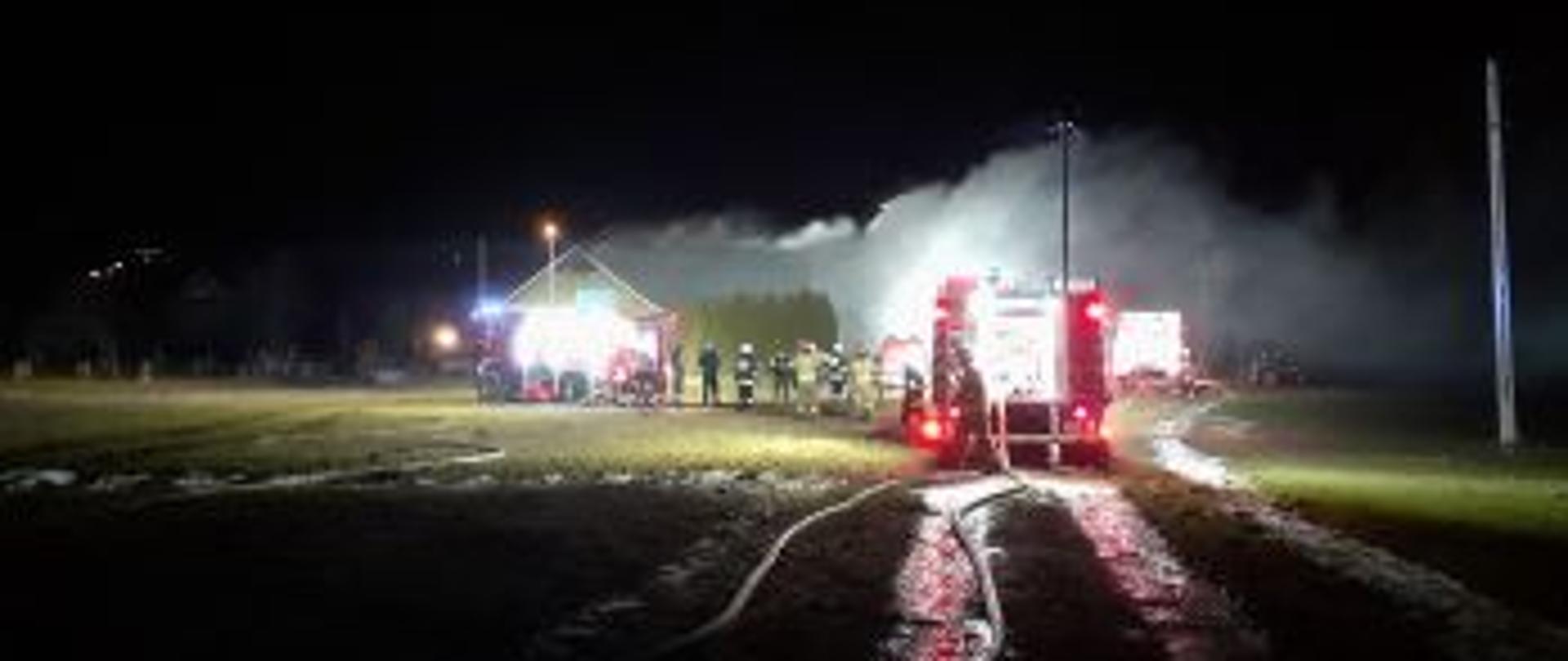 Zdjęcie przedstawia pożar garażu, stodoły i budynku mieszkalnego mającego miejsce w porze nocnej. W śrdkowej części zdjęcia widoczne są samochody strażackie. W oddali widoczna grupa strażaków. Z obiektów wydobywają się duże kłęby dymu.
