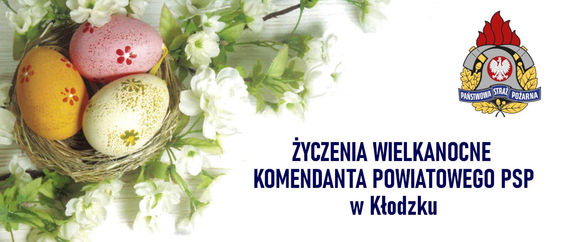 Baner wielkanocny z białym tłem. W górnym prawym rogu znajduje się godło PSP, a poniżej napis „Życzenia Wielkanocne Komendanta Powiatowego PSP w Kłodzku”. Po lewej stronie widoczne są wiosenne elementy: białe kwiaty oraz trzy kolorowe pisanki w gnieździe.