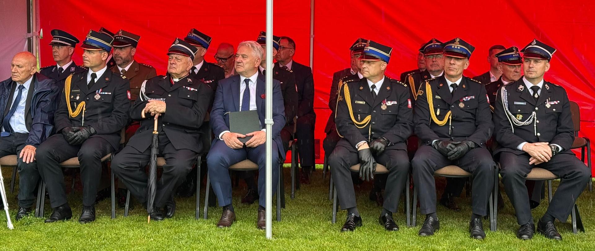 Zdjęcie przedstawia zaproszonych gości oraz wysokich rangą oficerów siedzących w rzędzie pod oficjalnym namiotem podczas uroczystości strażackiej. Zadaszenie stoiska: Nad siedzącymi rozciąga się intensywnie czerwony dach namiotu z dużym, białym, drukowanym napisem „STRAŻ POŻARNA”. Po prawej stronie widoczne jest logo Związku Ochotniczych Straży Pożarnych RP. Siedzący uczestnicy: W jednym rzędzie na krzesłach rozstawionych bezpośrednio na trawie siedzi kilkunastu mężczyzn. Większość z nich to oficerowie w ciemnogranatowych mundurach wyjściowych, czapkach rogatywkach ze złotymi galonami, w sznurach galowych i czarnych rękawiczkach. Ich piersi zdobią liczne baretki oraz medale .Goście cywilni: Wśród umundurowanych osób wyróżniają się dwaj mężczyźni w garniturach. Jeden (po lewej) ma na sobie ciemny garnitur i błękitną koszulę, drugi (bliżej środka) siedzi w niebieskim garniturze, białej koszuli i ciemnym krawacie, trzymając na kolanach ciemną teczkę z dokumentami. Konstrukcja: Przez środek kadru pionowo przebiega biała, metalowa rurka stelaża namiotu.