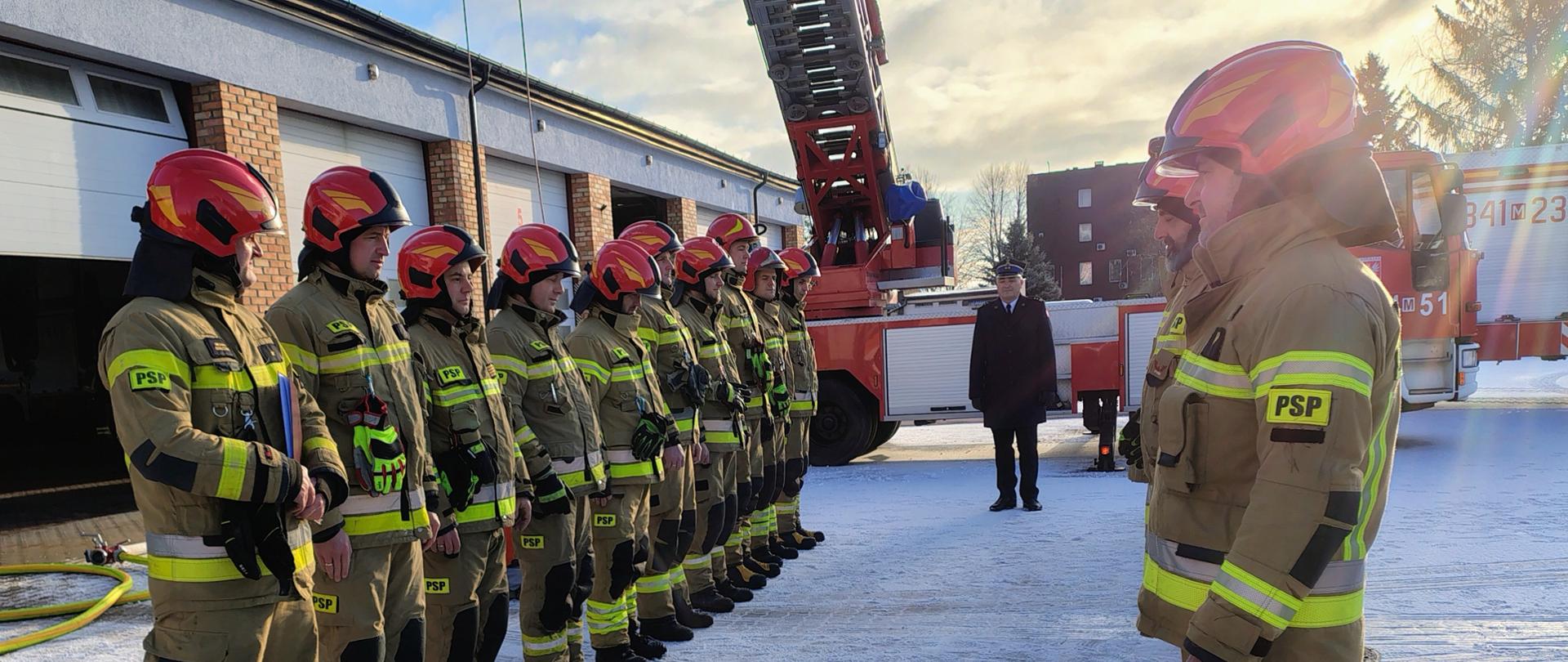 Grupa strażaków Państwowej Straży Pożarnej stoi w szeregu na ośnieżonym placu przed budynkiem garażowym jednostki podczas uroczystej zbiórki z okazji przejścia strażaka na zaopatrzenie emerytalne. Ubrani są w pełne umundurowanie bojowe z elementami odblaskowymi oraz czerwone hełmy. W tle widoczny jest samochód pożarniczy z wysuniętą drabiną mechaniczną oraz Komendant w płaszczu służbowym. Scena ma uroczysty, oficjalny charakter i odbywa się w zimowej scenerii, przy słonecznej pogodzie.