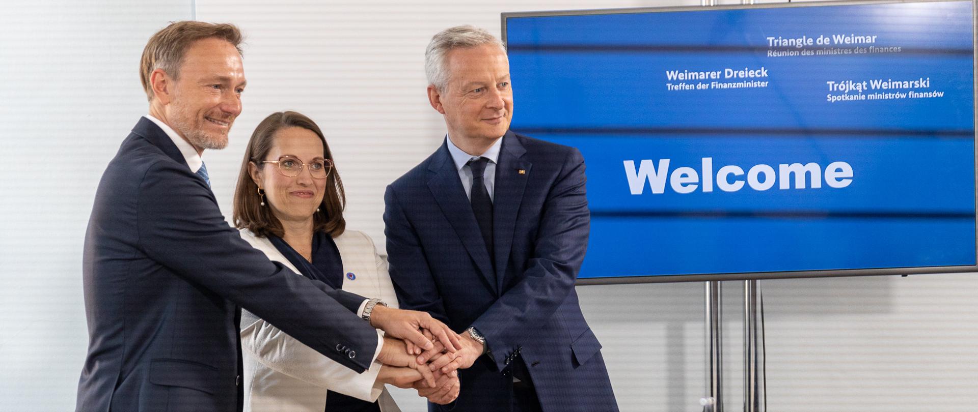 Minister Magdalena Rzeczkowska i ministrowie finansów Niemiec i Francji w powitalnym uścisku dłoni. Widoczny ekran z napisem „Welcome”