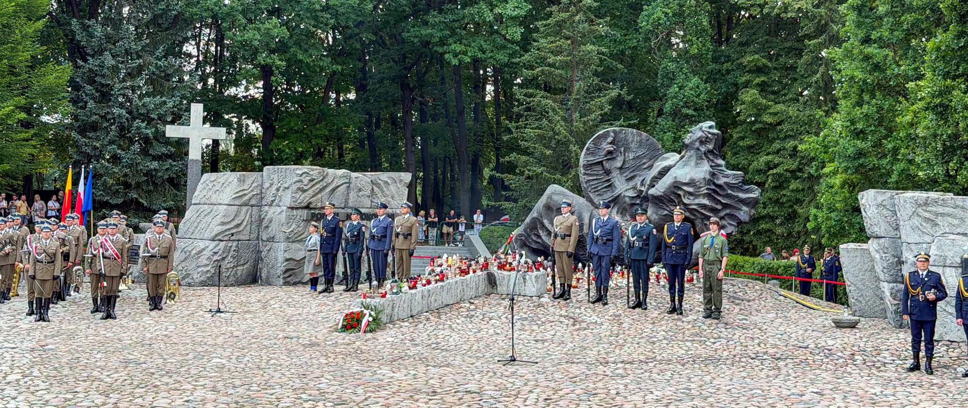 Uroczystość rocznicowa na cmentarzu. Przed pomnikiem stoją przedstawiciele służb mundurowych i poczty sztandarowe, a wokół monumentu płoną znicze.