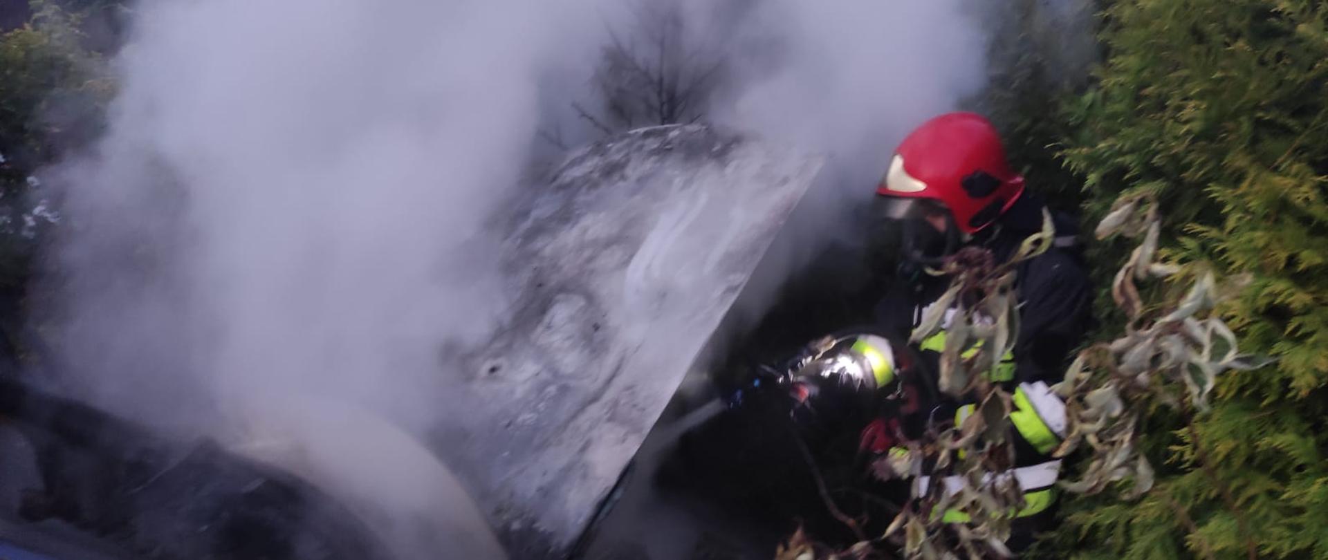 Zdjęcie przedstawia strażaka gaszącego komorę silnia w samochodzie osobowym, która objęta jest pożarem