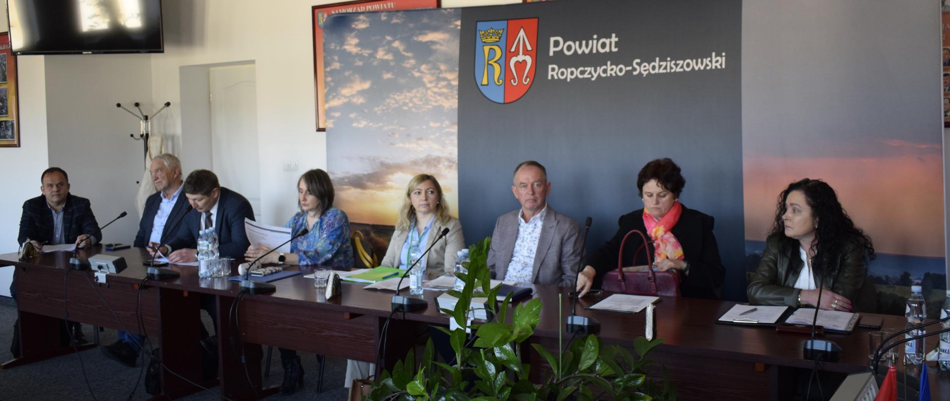 Grupa ludzi siedząca przy ciemnym stole. Cztery kobiety i czterech mężczyzn. Na stole leżą dokumenty, butelki z wodą i stoją mikrofony. Za ludźmi stoi duży ciemny baner z logiem. W lewym górnym rogu na ścianie wisi telewizor. Na pierwszym planie stoi zielony kwiatek.