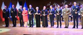 Szerokie ujęcie odznaczonych gości i funkcjonariuszy PSP stojących na scenie z nagrodami. W tle flagi narodowe i unijne oraz orkiestra.