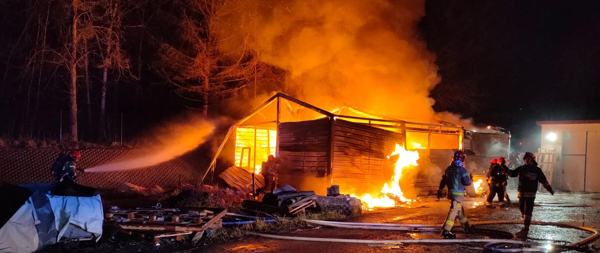 Na zdjęciu widać płonącą wiatę magazynową i strażaka, który prowadzi działania gaśnicze prądem wody. Płomienie wydostają się na zewnątrz. W tle widać przechodzących strażaków oraz inne zabudowania. 