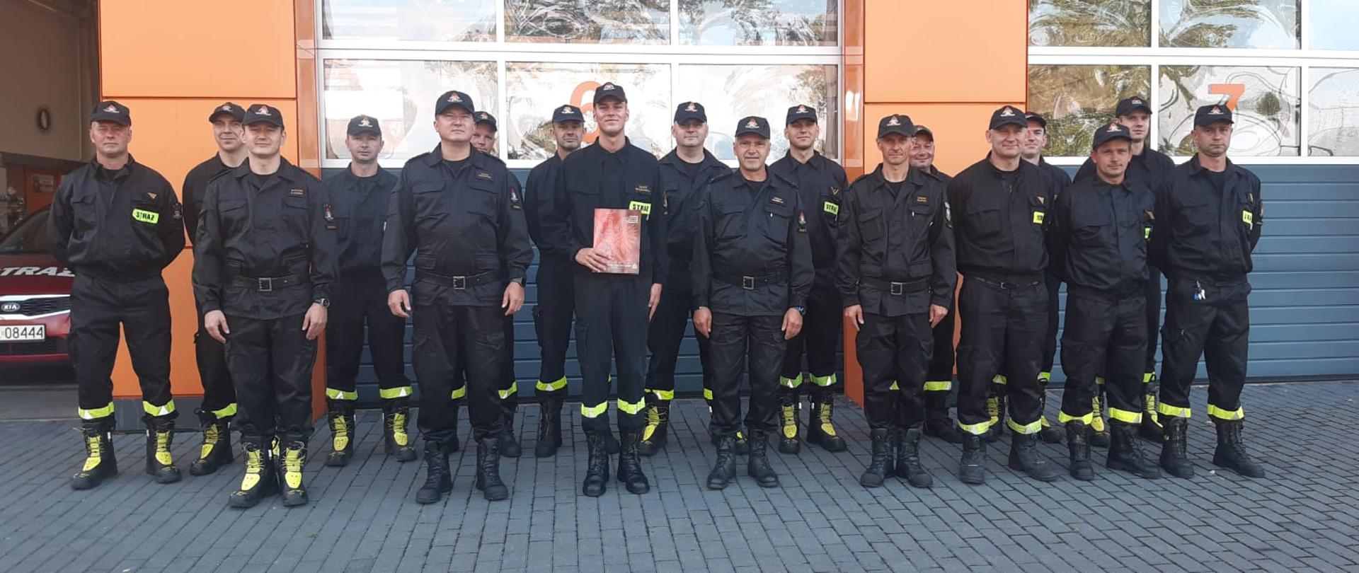 Na zdjęciu funkcjonariusze KP PSP w Wolsztynie, ubrani w czarne mundury. Stoją na wprost, zdjęcie grupowe. W tle bramy garażowe jednostki.