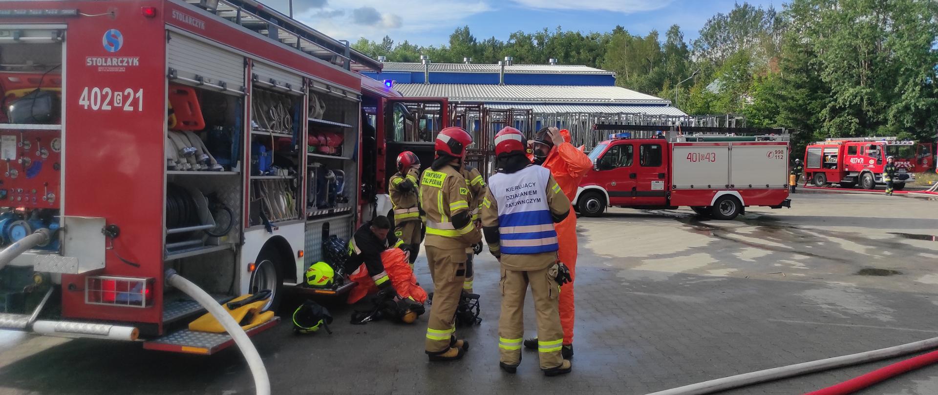 Z lewej strony zdjęcia przy wozie strażackim stoi grupa strażaków rozmawiających z mężczyzną w pomarańczowym kostiumie. Jeden z nich ma kamizelkę Kierujący Działaniem Ratowniczym. Jeden z strażaków, który siedzi na wozie, ubiera pomarańczowy kostium. Przed nimi stoją kolejne 2 wozy strażackie. Na ziemi leżą porozwijane węże strażackie