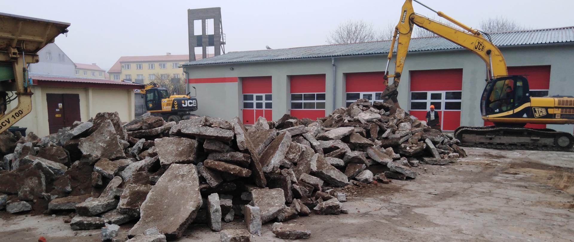 Zdjęcie przedstawiające na pierwszym planie usunięte płyty betonowe na tle wiat garażowych, koparki, obiekt energetyczny, oraz osobę. W tle budynki mieszkalne i korony drzew.