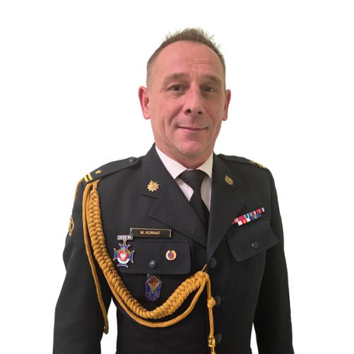 Zdjęcie przedstawia komendanta powiatowego Państwowej Straży Pożarnej w Łęczycy młodszego brygadiera Marcina Kornata w czarnym mundurze, na którym znajduje się stopień służbowy oraz odznaczenia strażackie. Zdjęcie portretowe na białym tle.