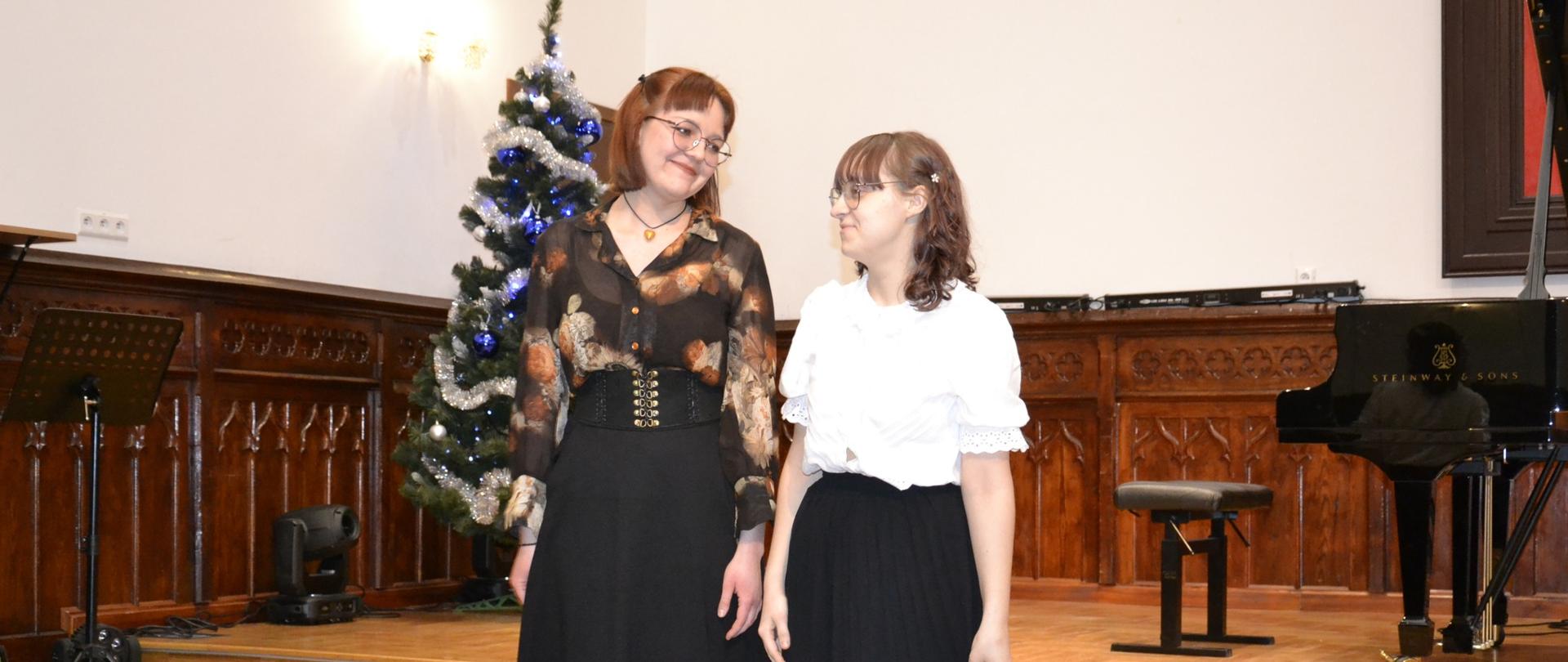 Dwie pianistki stoją na scenie w sali koncertowej przed publicznością, w tle widoczna jest choinka oraz fortepian.