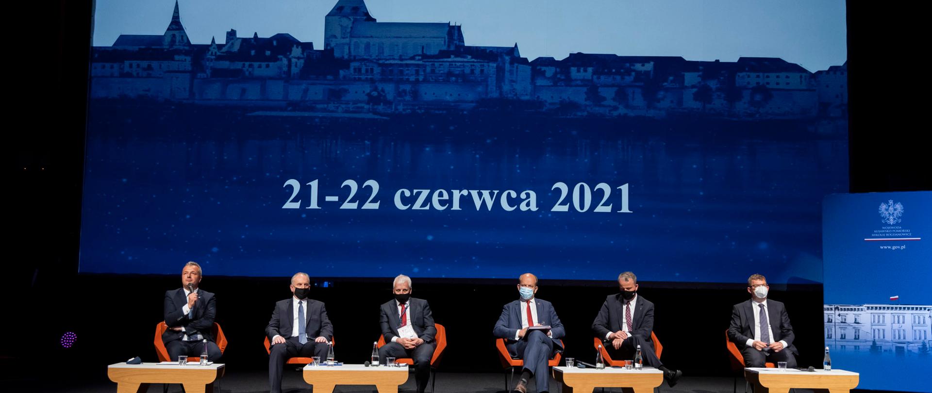 Wojewodowie Mikołaj Bogdanowicz, Wojewoda Kujawsko-Pomorski, Artur Chojecki, Wojewoda Warmińsko-Mazurski; Dariusz Drelich, Wojewoda Pomorski; Jarosław Obremski, Wojewoda Dolnośląski; Michał Zieliński, Wojewoda Wielkopolski siedzą w prezydium sali konferencyjnej. W tle widoczny ekran ze zdjęciem panoramy Torunia i napisem Welconomy 21-22 czerwca.
