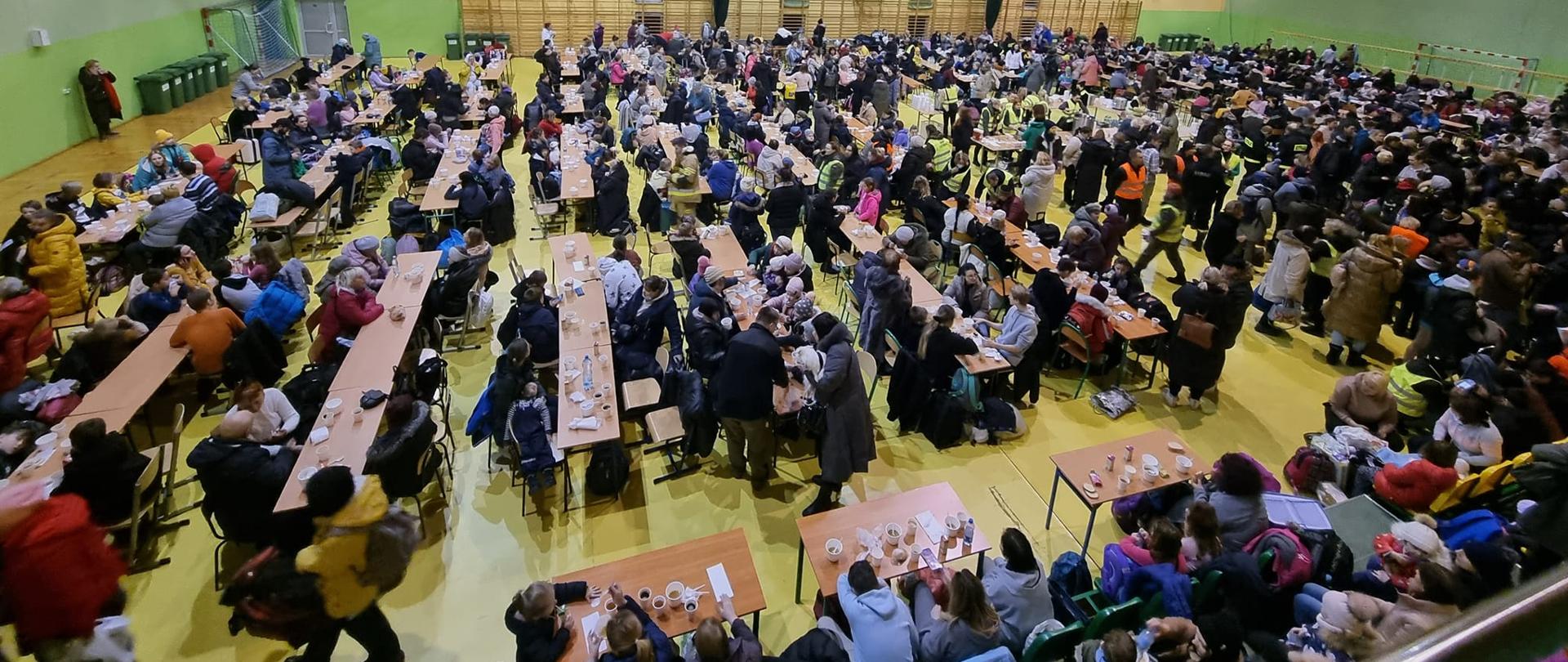 Zdjęcie przedstawia około 1000 uchodźców wojennych z Ukrainy, którzy znajdują się na hali sportowej Ośrodka Sportu i Rekreacji w Sędziszowie. Uchodźcy spożywają posiłki regeneracyjne przy stolikach.
