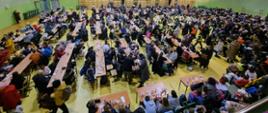 Zdjęcie przedstawia około 1000 uchodźców wojennych z Ukrainy, którzy znajdują się na hali sportowej Ośrodka Sportu i Rekreacji w Sędziszowie. Uchodźcy spożywają posiłki regeneracyjne przy stolikach.