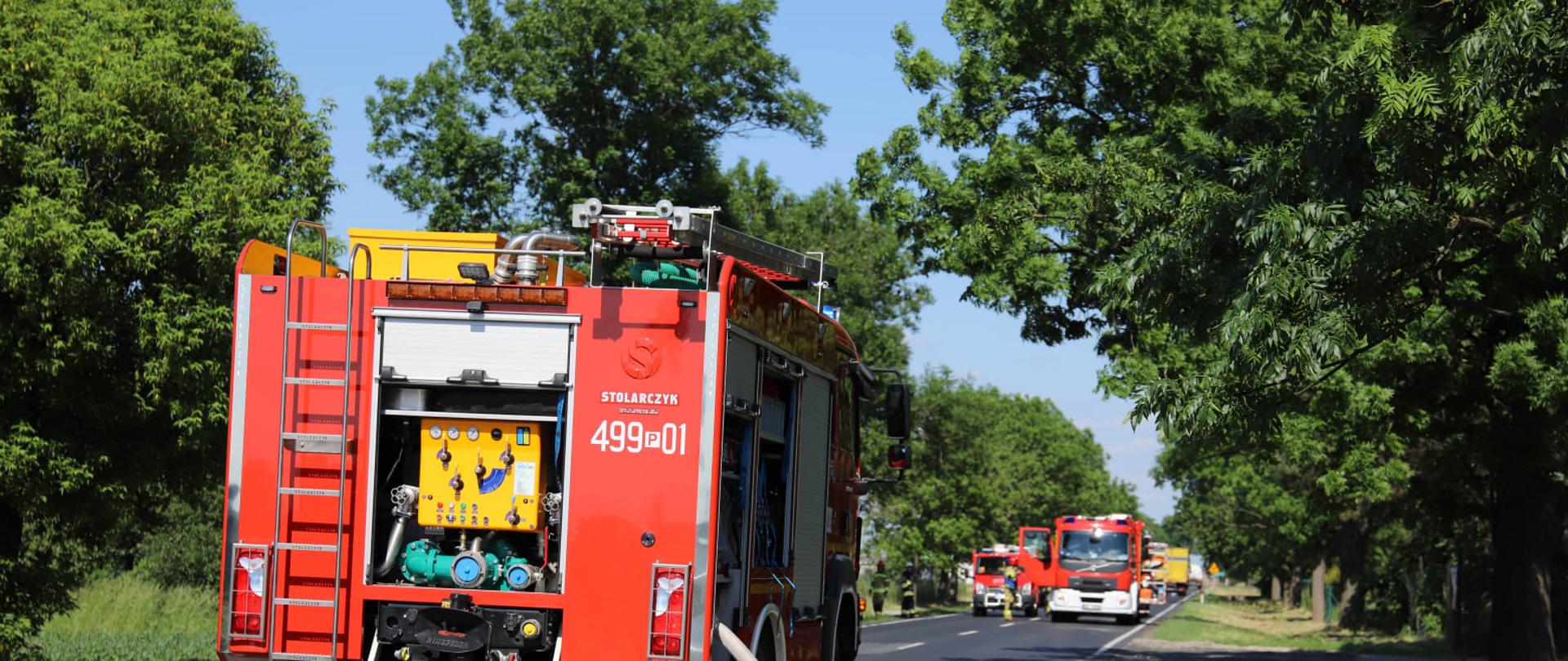 Na jezdni pojazdy pożarnicze blokujące drogę. Sprawiona linia gaśnicza celem zabezpieczenia pożarowego miejsca prowadzonych działań. Słoneczna pogoda, dużo zieleni wokoło 