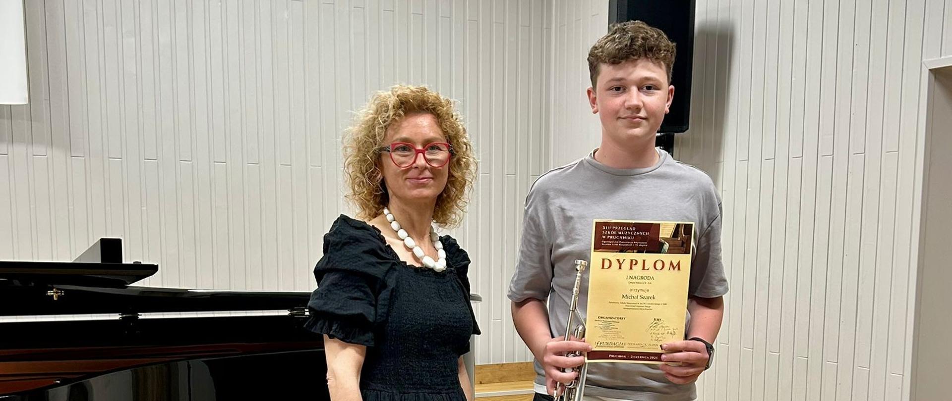 Uczeń stoi ze zdobytym dyplomem obok pani akompaniatorki. W tle widoczny jest fortepian, biała ściana oraz godło Polski.