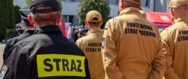 Funkcjonariusze Państwowej Straży Pożarnej stoją tyłem i słuchają apelu z okazji Dnia Strażaka