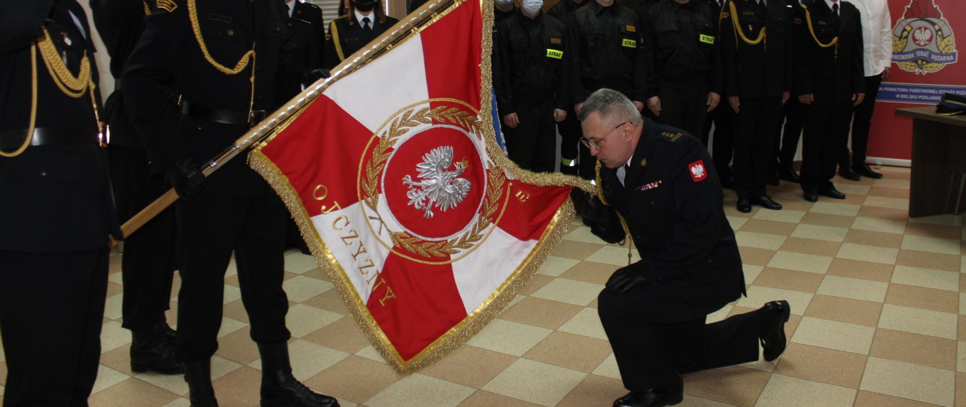 Zdjęcie przedstawia Komendanta Powiatowego PSP w Bielsku Podlaskim całującego sztandar komendy przyklękając na kolanie 