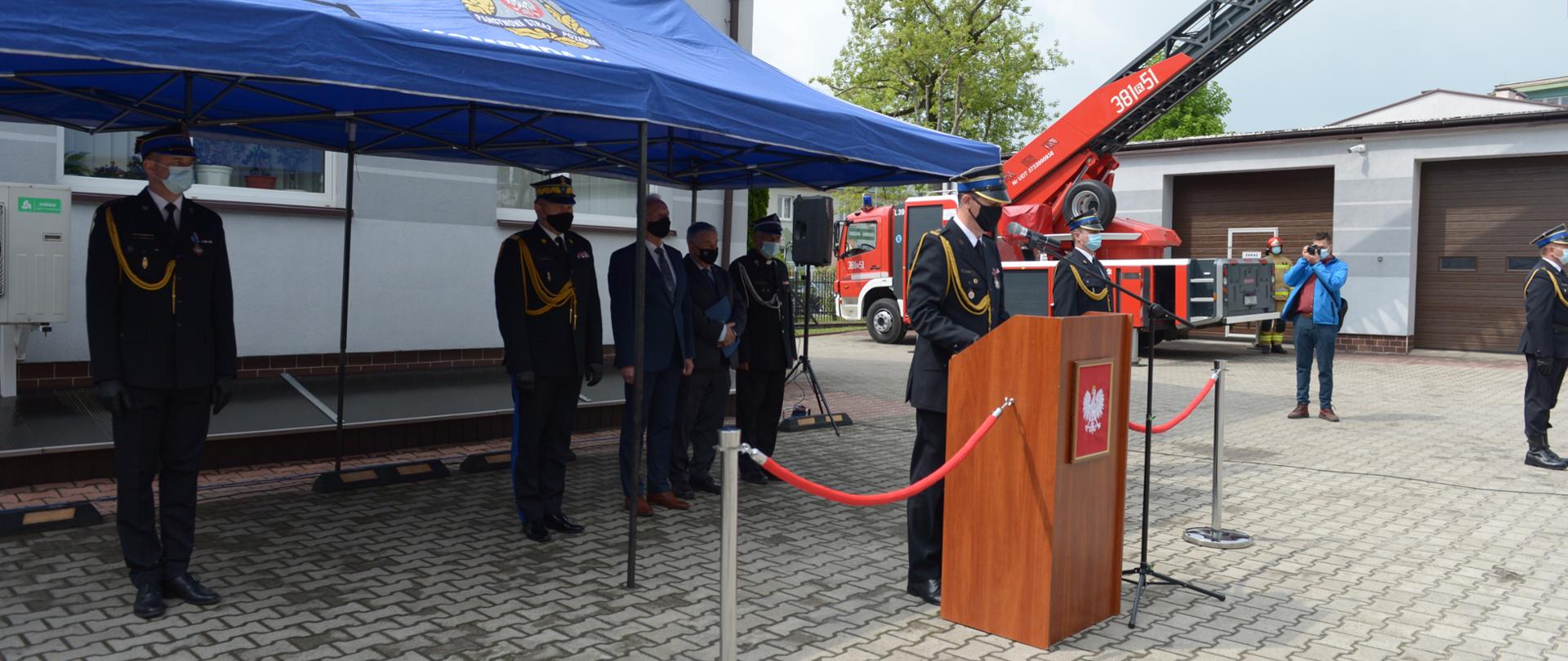 Przed budynkiem na placu komendy ustawiony zadaszony pawilon z logiem Państwowej Straży Pożarnej, pod którym stoi trzech strażaków w umundurowaniu wyjściowym oraz dwóch mężczyzn w garniturach. Przed pawilonem przy mównicy stoi strażak w umundurowaniu wyjściowym. W oddali widoczna wysunięta drabina samochodowa. Obok pawilonu stoi dwóch umundurowanych strażaków oraz mężczyzna, który wykonuje zdjęcie. 