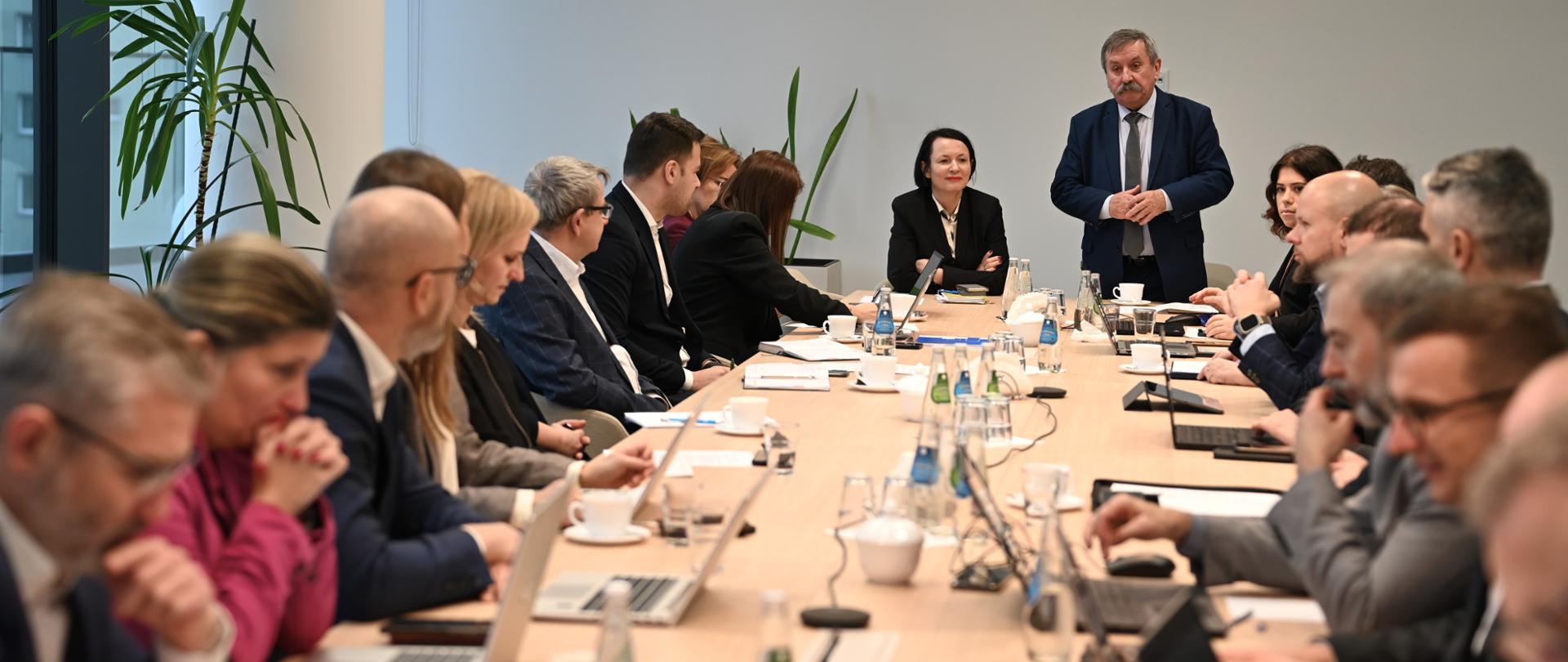 Zbliżenie na salę konferencyjną, gdzie Zastępca Prezesa Zarządu NFOŚiGW Józef Matysiak, ubrany w granatowy garnitur, stoi podczas przemówienia. Wokół drewnianego stołu siedzą uczestnicy spotkania, korzystający z laptopów i przeglądający dokumenty.