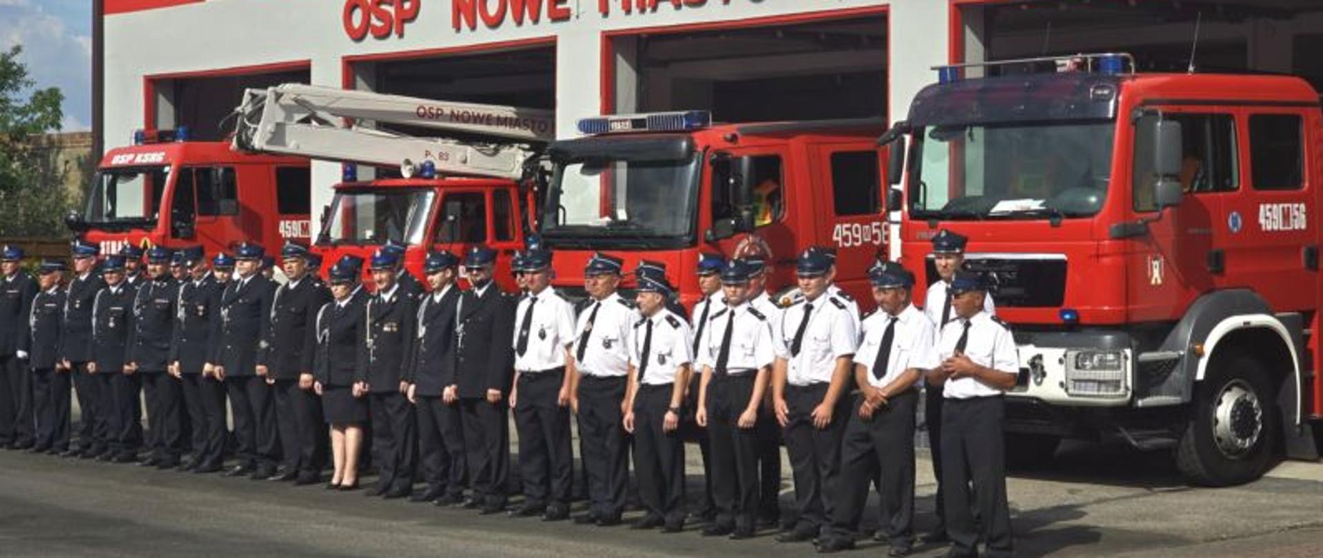 Grupa strażaków stoi w szeregu przed remizą OSP Nowe Miasto nad Pilicą. W tle znajdują się cztery czerwone wozy strażackie. Strażacy ubrani są w ciemne i jasne mundury – kobiety i mężczyźni stoją razem, patrząc w stronę fotografa. 