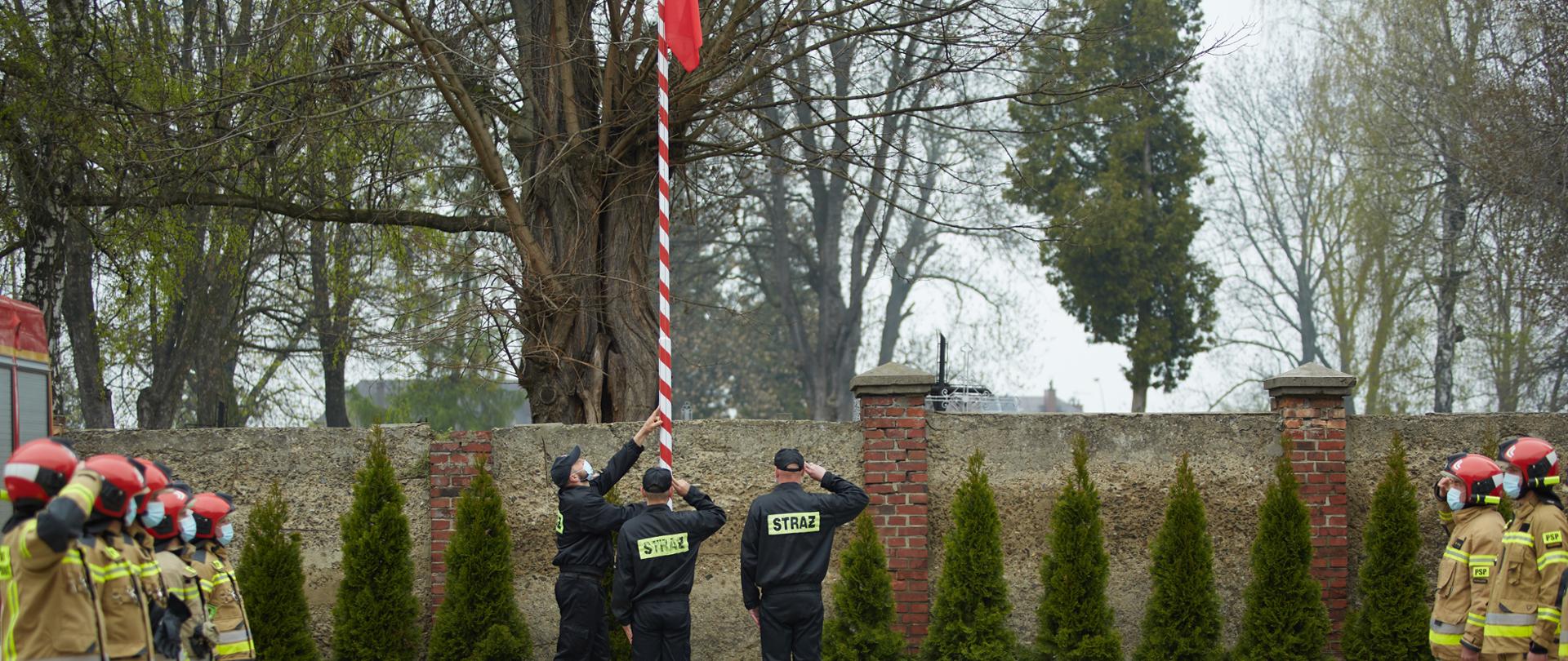 Na zdjęciu widoczna jest uroczysta zbiórka. Strażacy ubrani w mundury specjalne koloru żółtego stoją po lewej i prawej stronie w szeregach. W centralnym miejscu poczet flagowy składający się z trzech strażaków wciąga na maszt Flagę Polski. Dowódcy obu pododdziałów stojących po bokach salutują. 