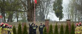 Na zdjęciu widoczna jest uroczysta zbiórka. Strażacy ubrani w mundury specjalne koloru żółtego stoją po lewej i prawej stronie w szeregach. W centralnym miejscu poczet flagowy składający się z trzech strażaków wciąga na maszt Flagę Polski. Dowódcy obu pododdziałów stojących po bokach salutują. 