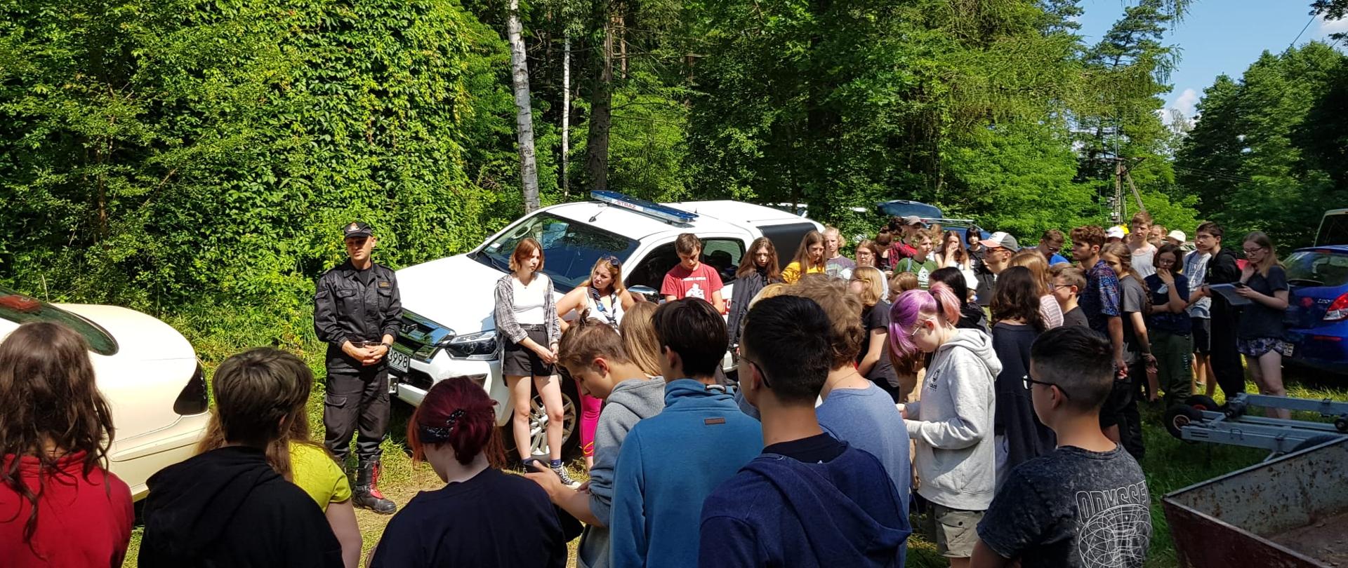Funkcjonariusz w mundurze dowódczo-sztabowym wygłasza pogadankę przed uczestnikami obozu harcerskiego. Dzieci i młodzież znajdują się na zewnątrz.