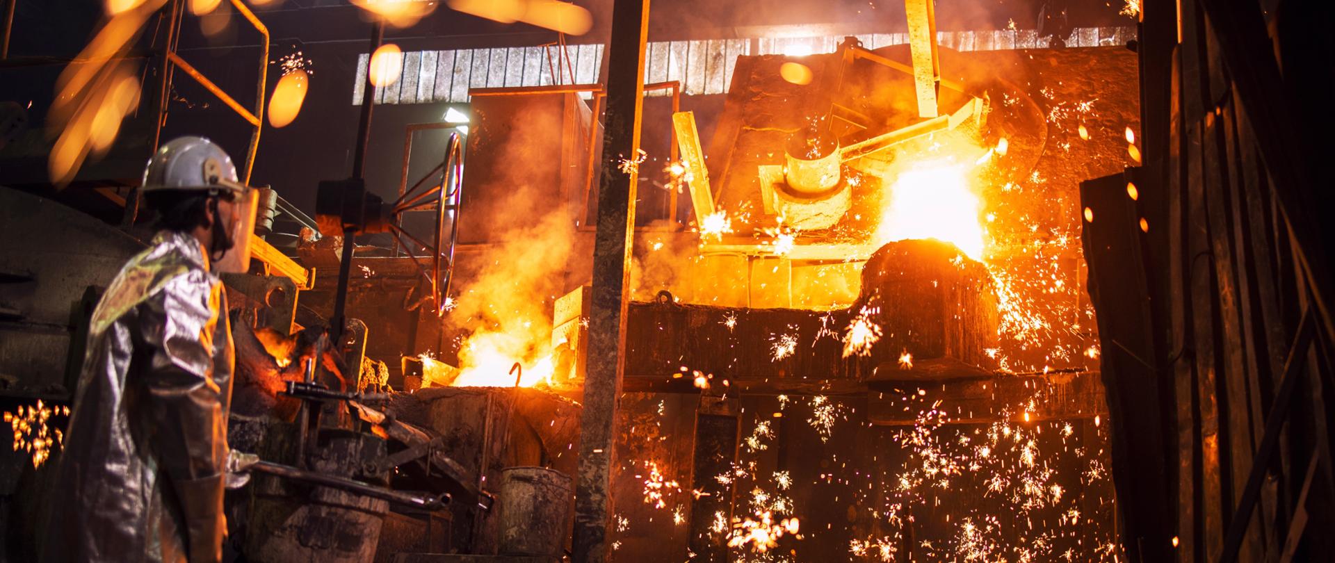 Po lewej stronie pracownik odlewni kontrolujący proces topienia żelaza w piecu. Lejąca się płonąca płynna stal i iskry latające dookoła. 