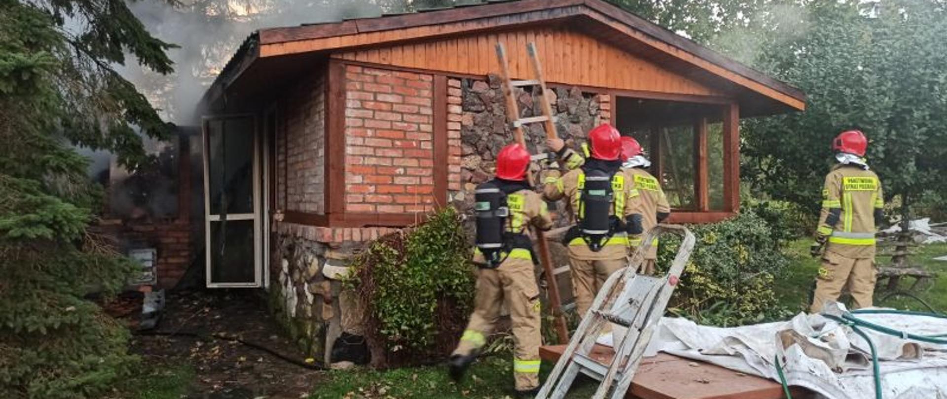 Na zdjęciu duża ogrodowa altanka. Budynek wykonany z czerwonej cegły i kamieni. Przy budynku stoi 4 strażaków, dwóch z nich ma na sobie aparaty ochrony dróg oddechowych. Strażacy stawiają drabinę przy budynku objętym pożarem. Z budynku wydostaje się dym