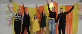 Grupa uczniów stoi na scenie przed kolorową, karnawałową dekoracją w odcieniach żółci i pomarańczu, unosząc ręce w górę w radosnym geście wspólnej zabawy.
