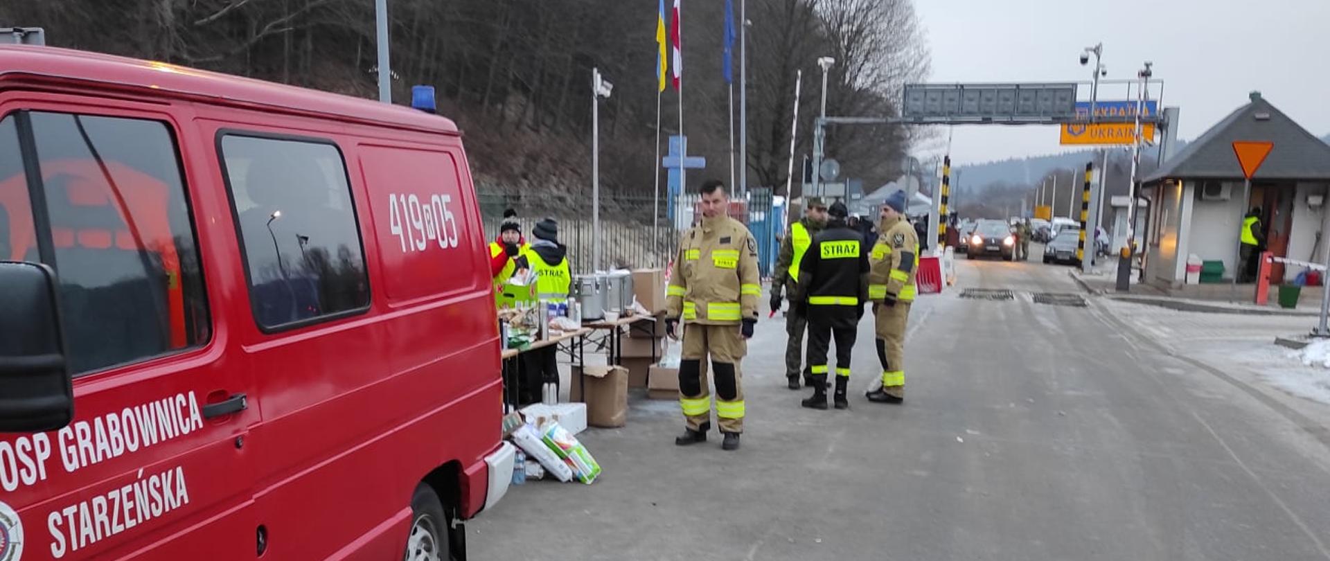 Strażacy OSP pomagają uchodźcom z Ukrainy. Fotografia wykonana w jasny dzień, widoczny czerwony samochód straży pożarnej i w tle kilku strażaków przy przejściu granicznym.