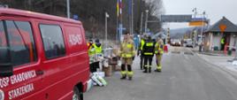 Strażacy OSP pomagają uchodźcom z Ukrainy. Fotografia wykonana w jasny dzień, widoczny czerwony samochód straży pożarnej i w tle kilku strażaków przy przejściu granicznym.