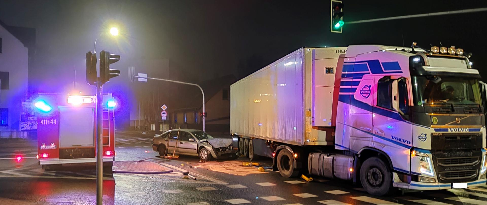Samochód ciężarowy z naczepą oraz uszkodzony samochód osobowy stojące po zderzeniu na jezdni. Obok strażacki samochód ratowniczo-gaśniczy.