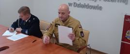 Przy stole siedzi komendant powiatowy Państwowej Straży Pożarnej w Działdowie oraz jeden ze strażaków-ochotników. Przeglądają kartki ze spisaną umową na pełnienie funkcji kierowcy w Ochotniczej Straży Pożarnej 