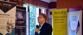 Mężczyzna w garniturze przemawia z mikrofonem na tle banerów reklamowych firm Mirion Technologies, GL Center, Revvity oraz Państwowej Inspekcji Sanitarne.