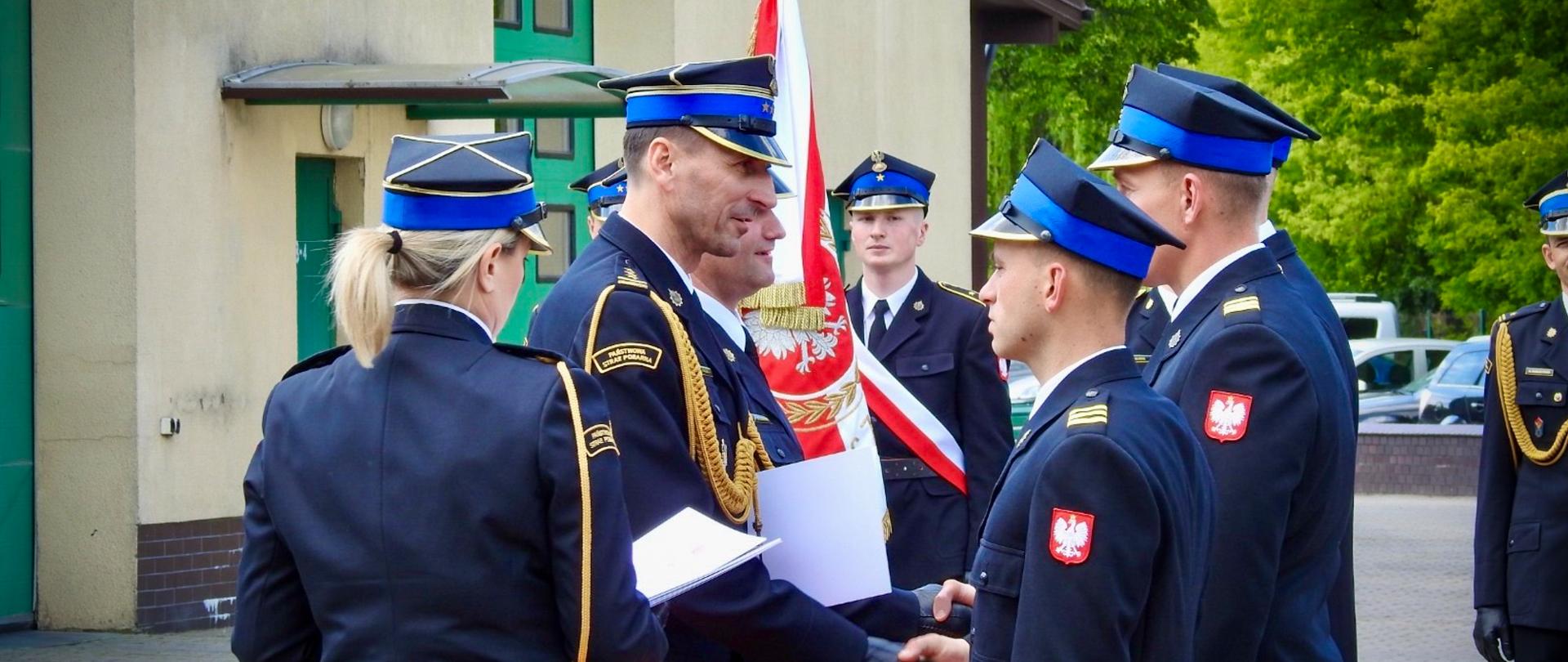 Łódzki Komendant wojewódzki PSP wręcza awans strażakowi ściskając mu dłoń i gratulując. W tle widać sztandar Komendy Powiatowej PSP w Tomaszowie Mazowieckim oraz innych strażaków