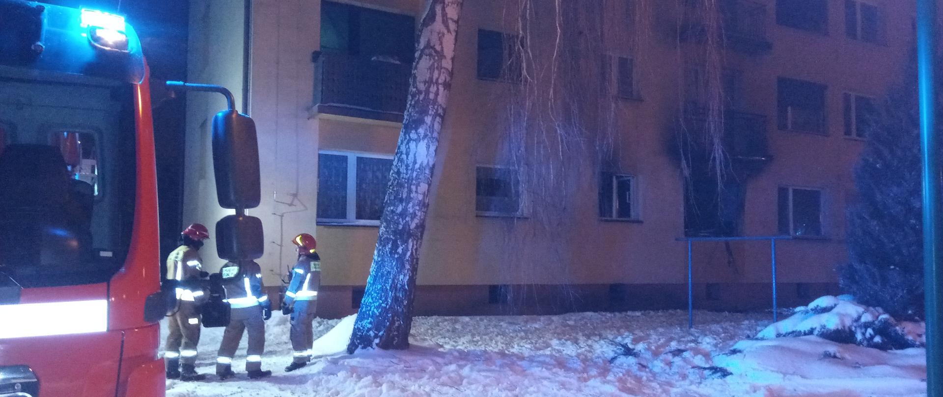 Zdjęcie zrobione na tle wielorodzinnego budynku mieszkalnego. Z lewej strony stoi samochód pożarniczy oraz trzech strażaków w umundurowaniu specjalnym, w hełmach. 