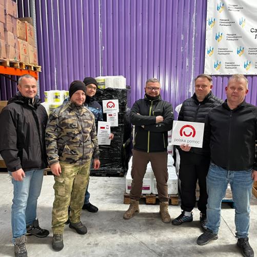 W magazynie na tle pudeł i palet z pomocą materialną do zdjęcia pozuje sześciu mężczyzn